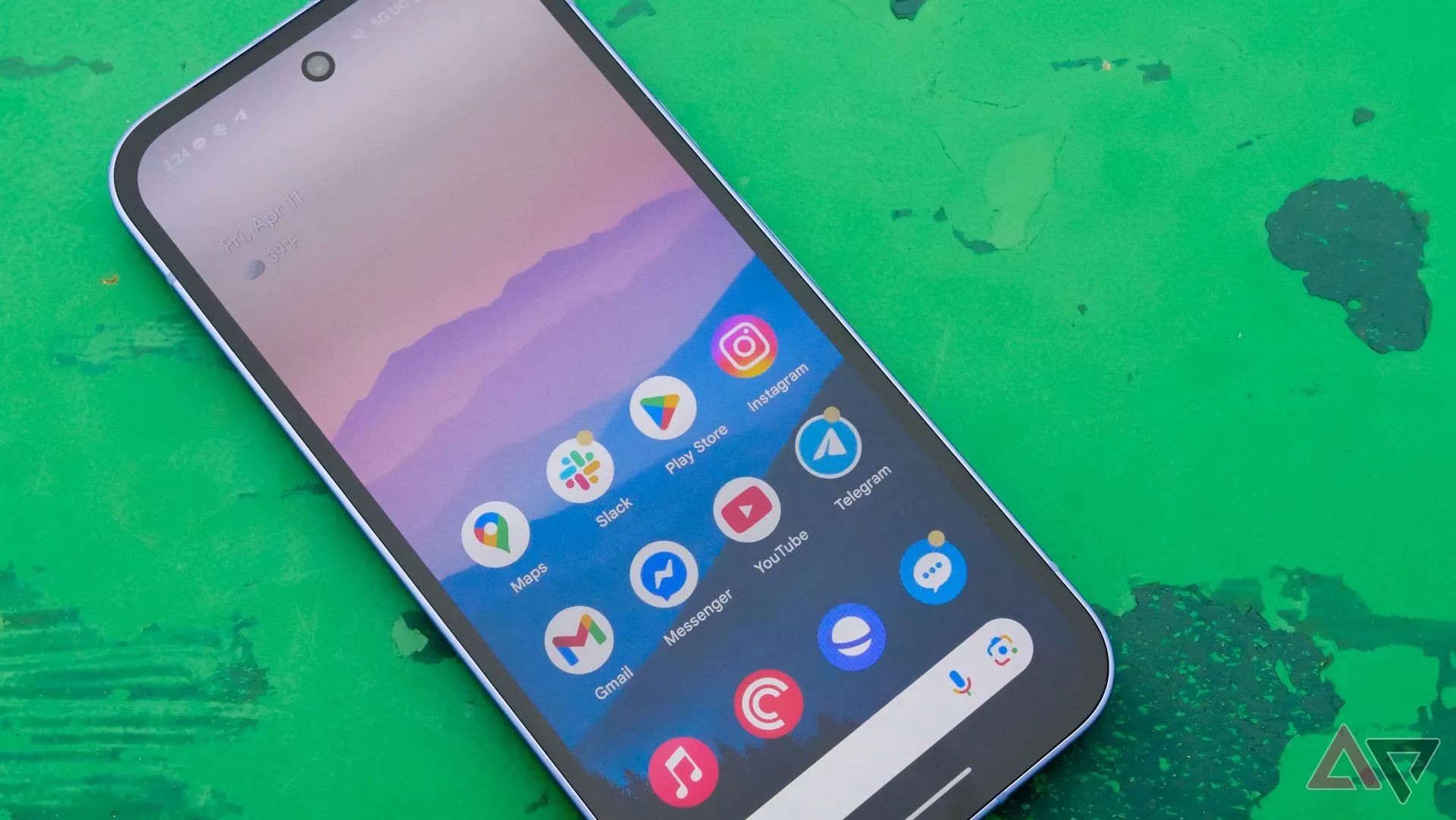 Google Pixel 9a màu xanh lá cây đặt trên bàn gỗ ngoài trời, màn hình hiển thị đang bật