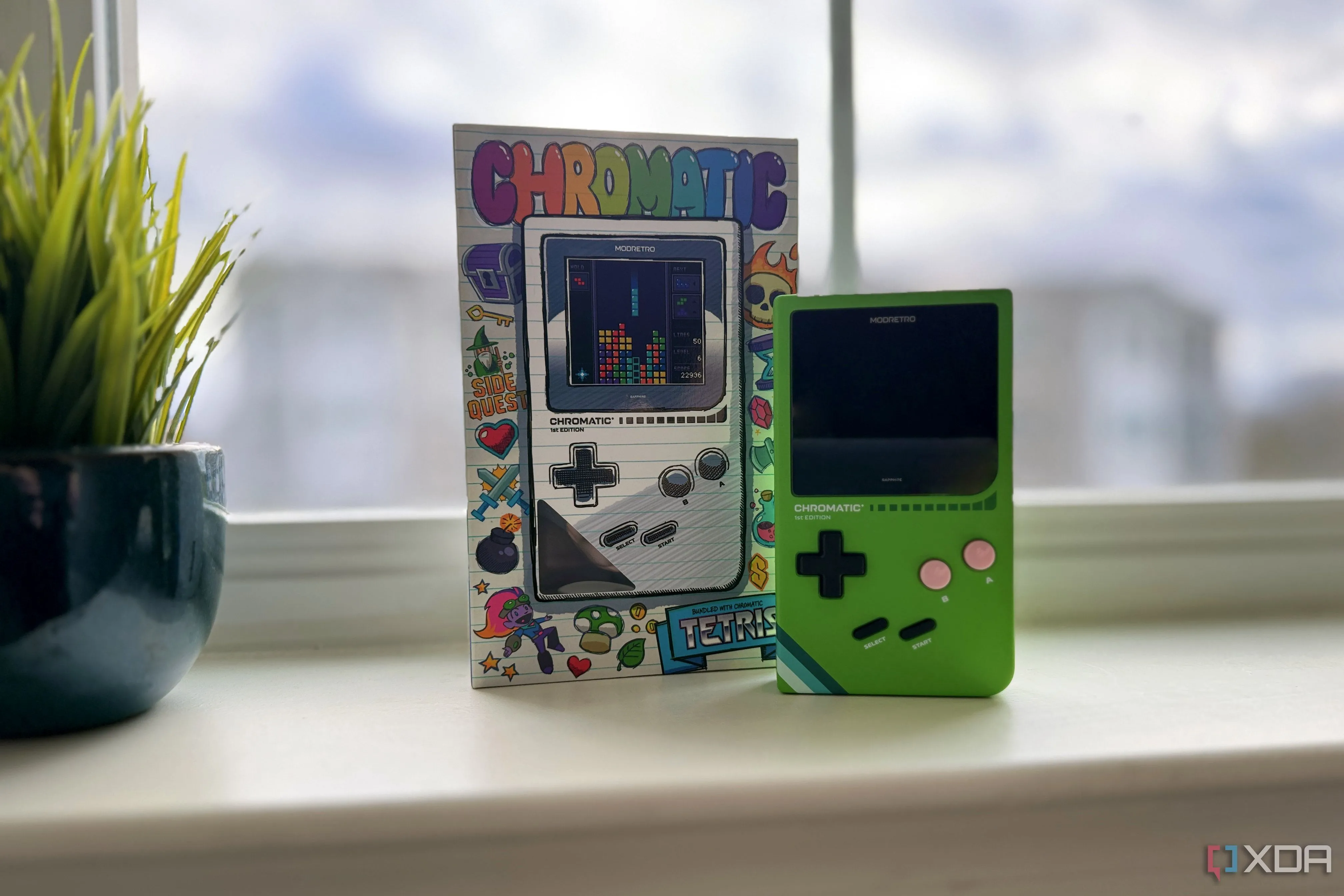 Máy chơi game ModRetro Chromatic bên cạnh hộp đựng trên bệ cửa sổ, biểu tượng cho game retro và console mod